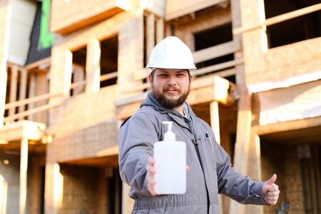 Builder showing disinfectant at construction site and wearing hardhat, building in background. Concept of coronavirus protection.の写真素材