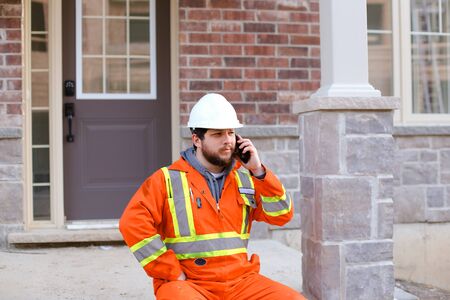 Caucasian foreman sitting in front of house and talkign by smartphone, wearing orange workwear. Concept of modern technology and builder profession.の写真素材