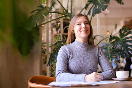 Young female student taking notes into notebook in class room, monstera in background and cup of tea on table. Concept of university life and interesting lesson.の写真素材
