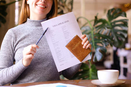 Young smiling caucasian woman filling visa application and holding passport at cabinet, monstera i background. Concept of travel agency and execution of documents.の写真素材