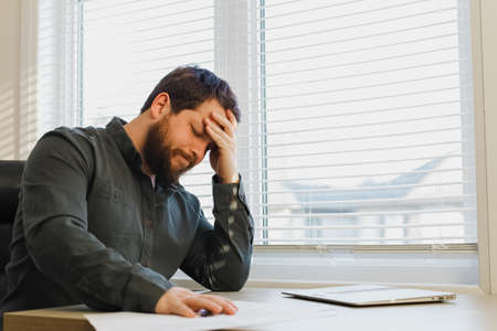 Tired thinking boss working with documents at cabinet near window, closed laptop on desk. Concept of financial crisis because of coronavirus.の写真素材