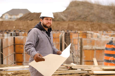 Male caucasian engineer holding drawing plan of hause at construction site, wearing white hardhat. Concept of hause building and builder profession.の写真素材