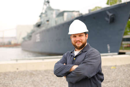 Portrait of marine first mate standing near big vessel in background and wearing helmet with work jumpsuit. Concept of maritime job and profession and marine team.の写真素材