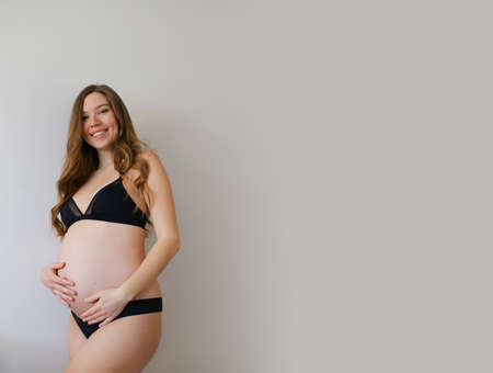 Beautiful pregnant american woman in black lingerie holding her belly in grey copy space background. Concept of expectant female person pohto session and maternity.の写真素材