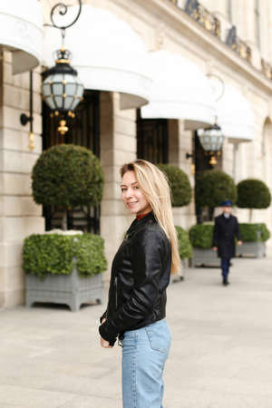 Young blonde woman in leather jacket walking in city and standing near building. Concept of fashionable look and walk in european town.の写真素材