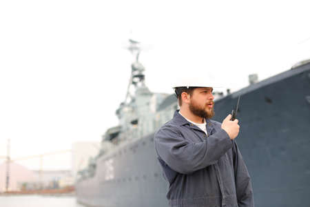 Captain wearing helmet and talking by VHF walkie talkie radio, standing on coast with offshore vessel in background. Concept of maritime job and engineering department of marine team.の写真素材