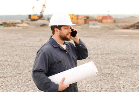 American foreman speaking by VHF walkie talkie and blueprints papers on construction site, wearing helmet and work jumpsuit. Concept of builder profession.の写真素材