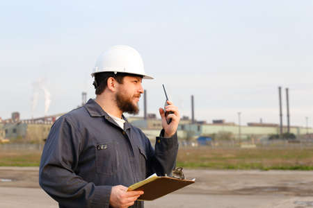Portrait of builder engineer standing on construction site and holding papers. Man wearing white helmet and work jumpsuit. Concept of industry profession and building engineering job.の写真素材