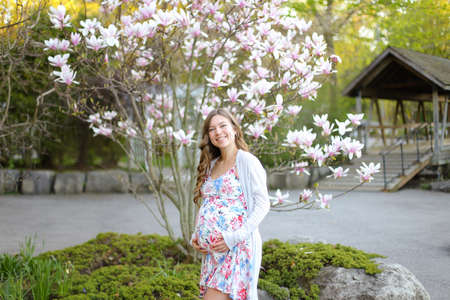 Young pregnant woman standing near blooming tree in park. Female person wearing dress. Concept of maternity, motherhood and springtime.の写真素材