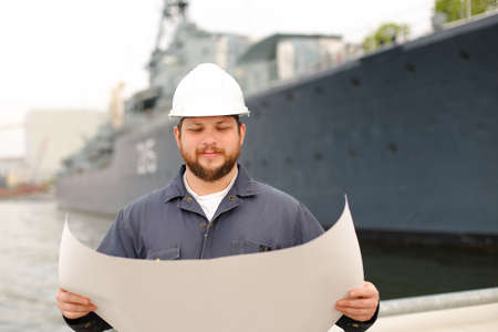 Assistant engineer wearing helmet and holding blueprints, standing on coast near vessel. Concept of...