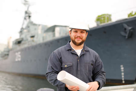 Marine assistant engineer wearing helmet and holding blueprints, standing on coast near vessel. Concept of maritime job and engineering department.の写真素材