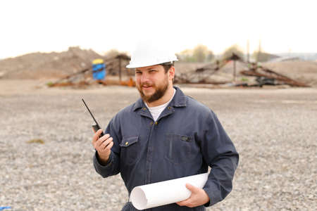 Portrait of caucasian foreman standing on construction site, talking by VHF walkie talkie, wearing helmet, work jumpsuit and holding blueprints. Concept of urban works.の写真素材