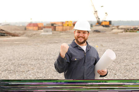 Happy male foreman holding blueprints papers on construction site, wearing helmet and work jumpsuit....