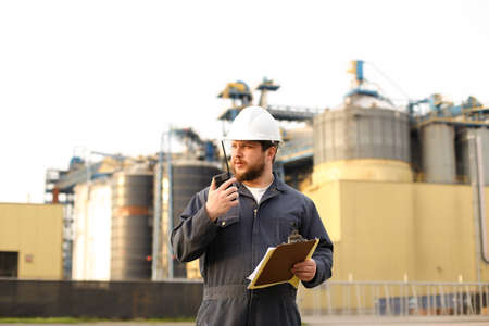 American engineer holding VHF walkie talkie and notes, factory in background. Man wearing helmet and work jumpsuit. Concept of engineering profession and modern technology, worker.の写真素材