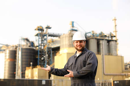 Portrait of male industrial director and engineer showing factory exterior outdoors. Man wearing white helmet and blue work jumsuit. Concept of plant jobs, engineering profession.の写真素材