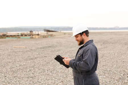 Foreman in helmet surfing internet by tablet on construction site, wearing jumpsuit. Concept of wireless modern technology and building engineer.の写真素材