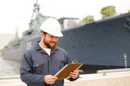 Deckhand wearing helmet and holding papers documents, standing on coast near vessel. Concept of maritime job and engineering department of marine team.の写真素材