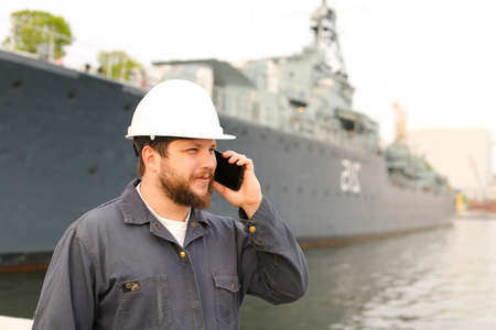 Marine captain speaking by VHF walkie talkie near vessel in background, wearing helmet and work jumpsuit. Concept of maritime profession , job and seaman.の写真素材