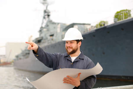 Marine engineer wearing helmet and holding blueprints, showing forefinger and standing on coast near vessel. Concept of maritime job and engineering department.の写真素材