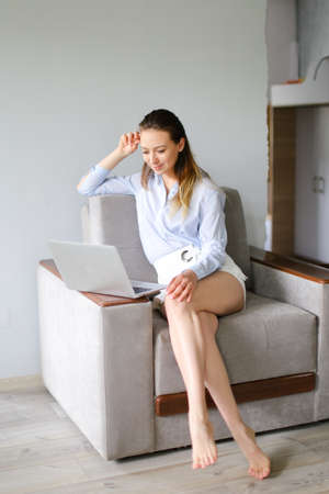 Young female freelancer working with laptop at home and sitting on sofa. Caucasian woman surfing internet and wearing white shirt. Concept of frelance job, guarantine time and modern technology.の写真素材