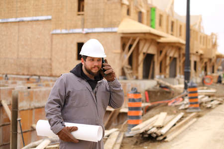 Foreman speaking by mobile phone and holding drawing plan at construction site, wearing hardhat. Concept of architectural profession and house building.の写真素材