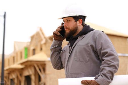 Caucasian builder talking by mobile phone and holding drawing plan at construction site, wearing hardhat. Concept of architectural profession and house building.の写真素材