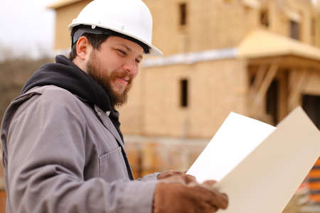 Portrait of male foreman holding drawing plan of hause at construction site, wearing white hardhat. Concept of hause building and builder profession.の写真素材