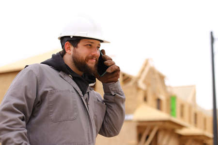 Caucasian builder talking by mobile phone at construction site, wearing hardhat. Concept of architectural profession and house building.の写真素材