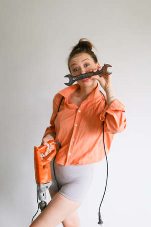Young woman holding a drill and kissing wrench, wearing orange shirt standing on isolated background. Concept of repairs tools and attractive wife.の写真素材