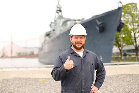 Portrait of marine captain standing near big vessel in background, showing thumbs up and wearing helmet with work jumpsuit. Concept of maritime job and profession, marine team.の写真素材