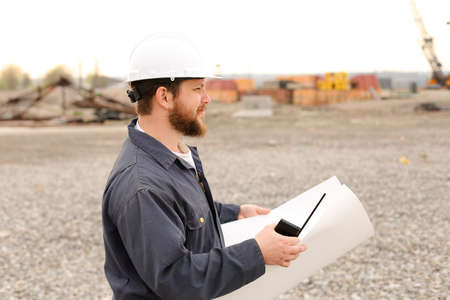 Foreman holding VHF walkie talkie and blueprints papers on construction site, wearing helmet and work jumpsuit. Concept of builde profession.の写真素材