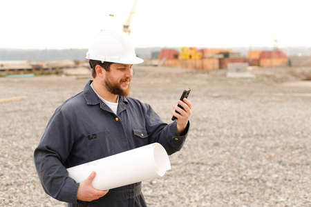 American building enginer holding VHF walkie talkie and blueprints papers on construction site, wearing helmet and work jumpsuit. Concept of builder profession.の写真素材