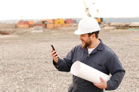 American male building enginer holding VHF walkie talkie and blueprints papers on construction site, wearing helmet and work jumpsuit. Concept of builder profession.の写真素材