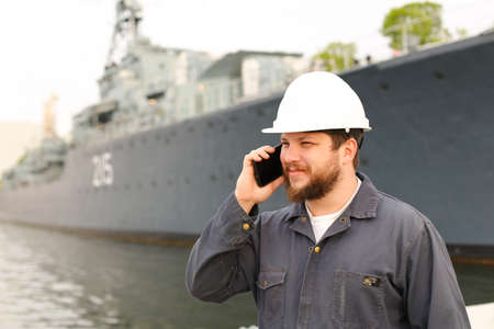 Marine deck officer speaking by VHF walkie talkie near vessel in background, wearing helmet and work jumpsuit. Concept of maritime profession , job and seaman.の写真素材