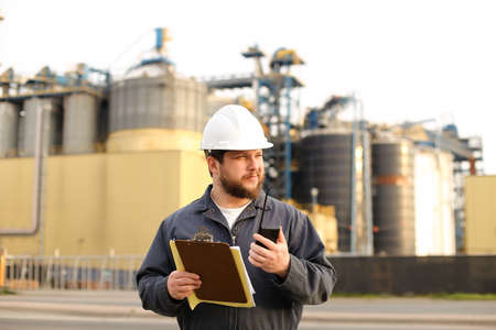 Caucasian engineer holding VHF walkie talkie and notes, factory in background. Man wearing helmet...