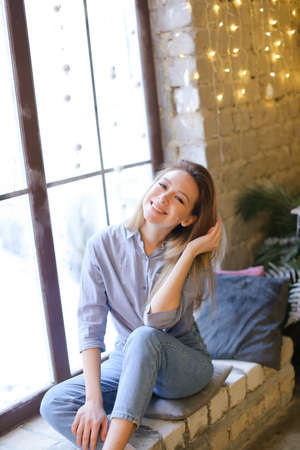 Smiling blonde girl sitting on windowsill near decorated window by garlands.の写真素材