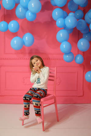Little girl sitting on chair near many balloons, pink wall in background.の写真素材