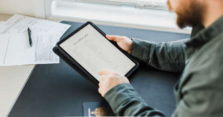 Man getting visa and reading application on tablet, passport on desk.