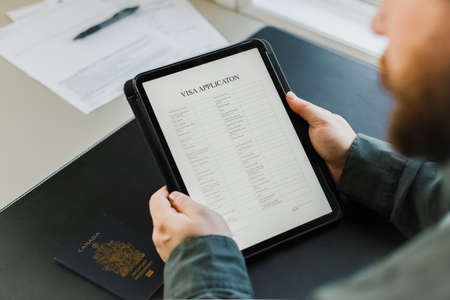 Close up bearded man getting visa and reading application on tablet, passport on desk.の写真素材
