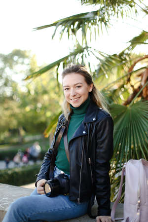 Young woman sitting with camera outside near palm leaves.