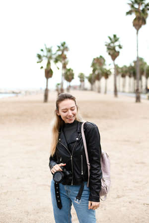 Young woman walking on beach with camera in leather jacket.の写真素材