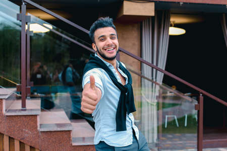 Cheerful young man looks Muslim smiling and showing hand gesture Cool , Is next to restaurant on background of stairs outdoors.の写真素材
