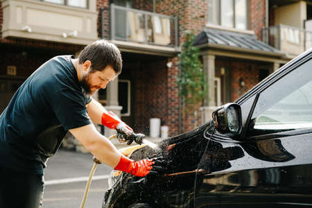 European man washing car by water and jet in front of house.