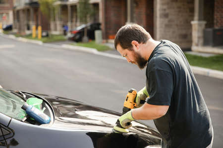 Caucasian bearded man polishing car by polisher in front of house.の写真素材