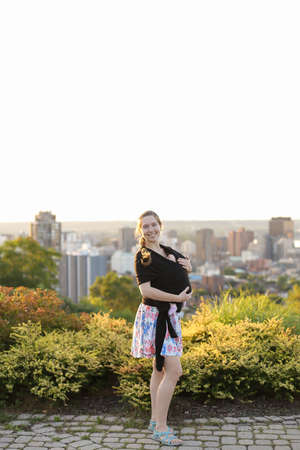 Caucasian mother carrying babe in sling city in background.の写真素材