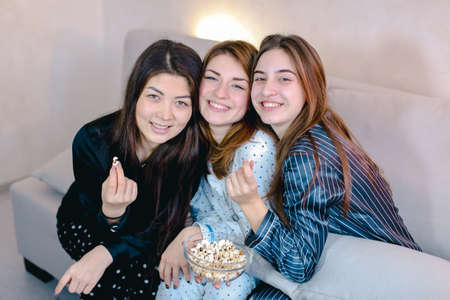 Friendly female friends cool spend time and eat popcorn, sitting on soft sofa in bright bedroom in evening.の写真素材