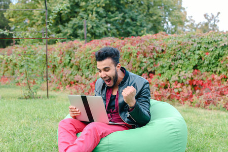 Successful Arabic Young Businessman Sits With Laptop in Chair, Stares at Laptop Screen, Raises Hands, Making Hand Gesture yes in Garden Outdoors.の写真素材