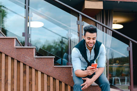 Attractive young Arabic man standing with suitcase in hand and posing for on camera, put one foot before the other and is based on the stairs near the restaurant outdoors.の写真素材