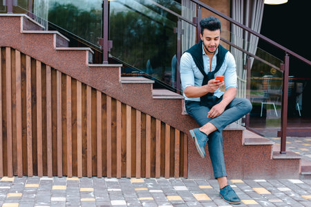 Attractive young Arabic man standing with suitcase in hand and posing for on camera, put one foot before the other and is based on the stairs near the restaurant outdoors.の写真素材