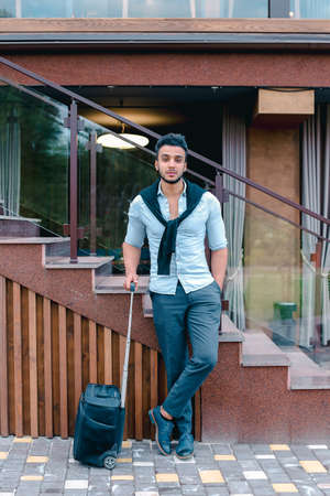 Attractive young Arabic man standing with suitcase in hand and posing for on camera, put one foot before the other and is based on the stairs near the restaurant outdoors.の写真素材